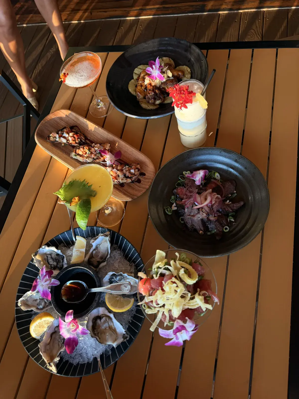 A full table of pupus at Voyagers in Kailua-Kona at golden hour: oysters on ice, lomi salmon bruschetta, hulihuli chicken wings, ahi poke parfait, sliced steak and tropical cocktails