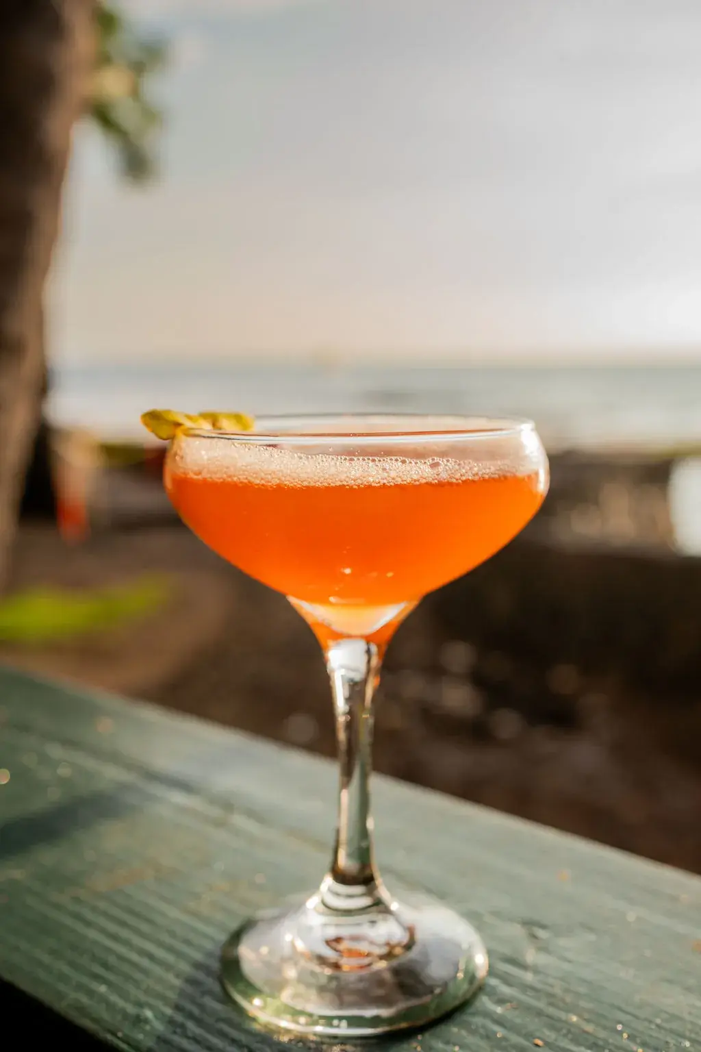A coupe cocktail on the Voyagers rail at golden hour with lava rock and the Kona coast behind it